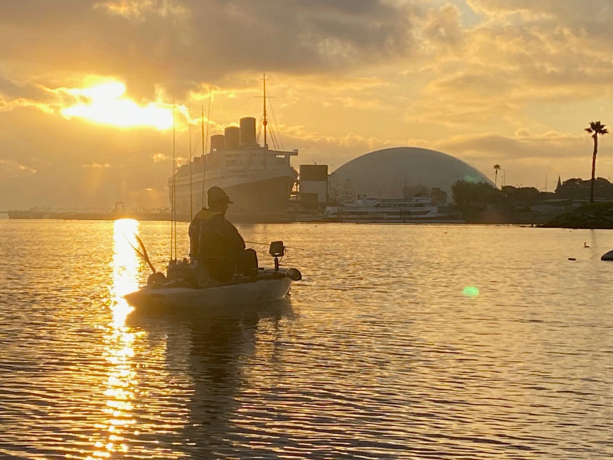Person fishing on a small boat at sunset with a large ship and dome-shaped building in the background.